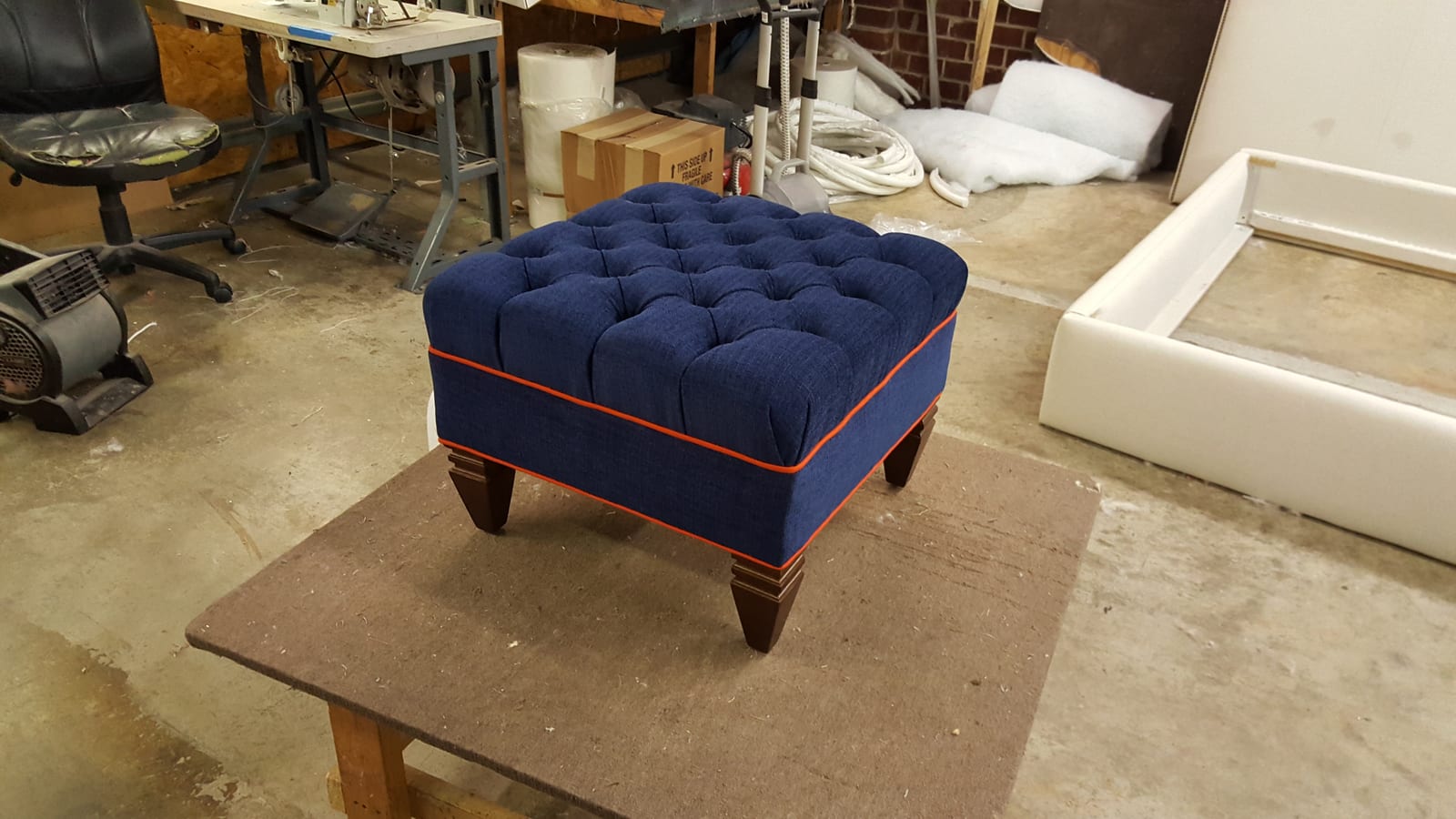 A navy tufted ottoman with orange contrast piping and wood legs